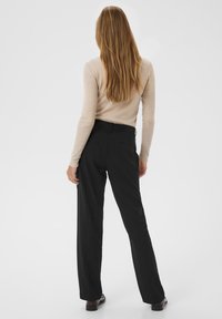 Femme aux longs cheveux auburn se tenant de dos, portant un haut beige à manches longues et un pantalon noir à jambes droites avec des chaussures sombres.