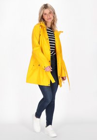 Rumena nepremočljiva dežna jakna s kapuco, s snemljivimi gumbi in stranskimi žepi, oblečena čez črno-belo črtasto majico in modre kavbojke.