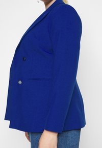 Blauwe dubbele boord blazer met een getailleerde pasvorm, gemaakt van een gladde stof. Voorzien van twee knopen en een enkele borstzak.