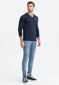 T-paita, jossa on pitkät hihat ja V-kaulus, valmistettu pehmeästä materiaalista, yhdistetty vaaleansinisiin istuviin farkkuihin ja tummansinisiin tennareihin, joissa on valkoiset nauhat ja yksityiskohdat.