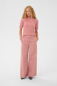 Kvinde med krøllet blont hår iført en pink ribbet kortærmet turtleneck og brede pink bukser, stående med hænderne i lommerne.