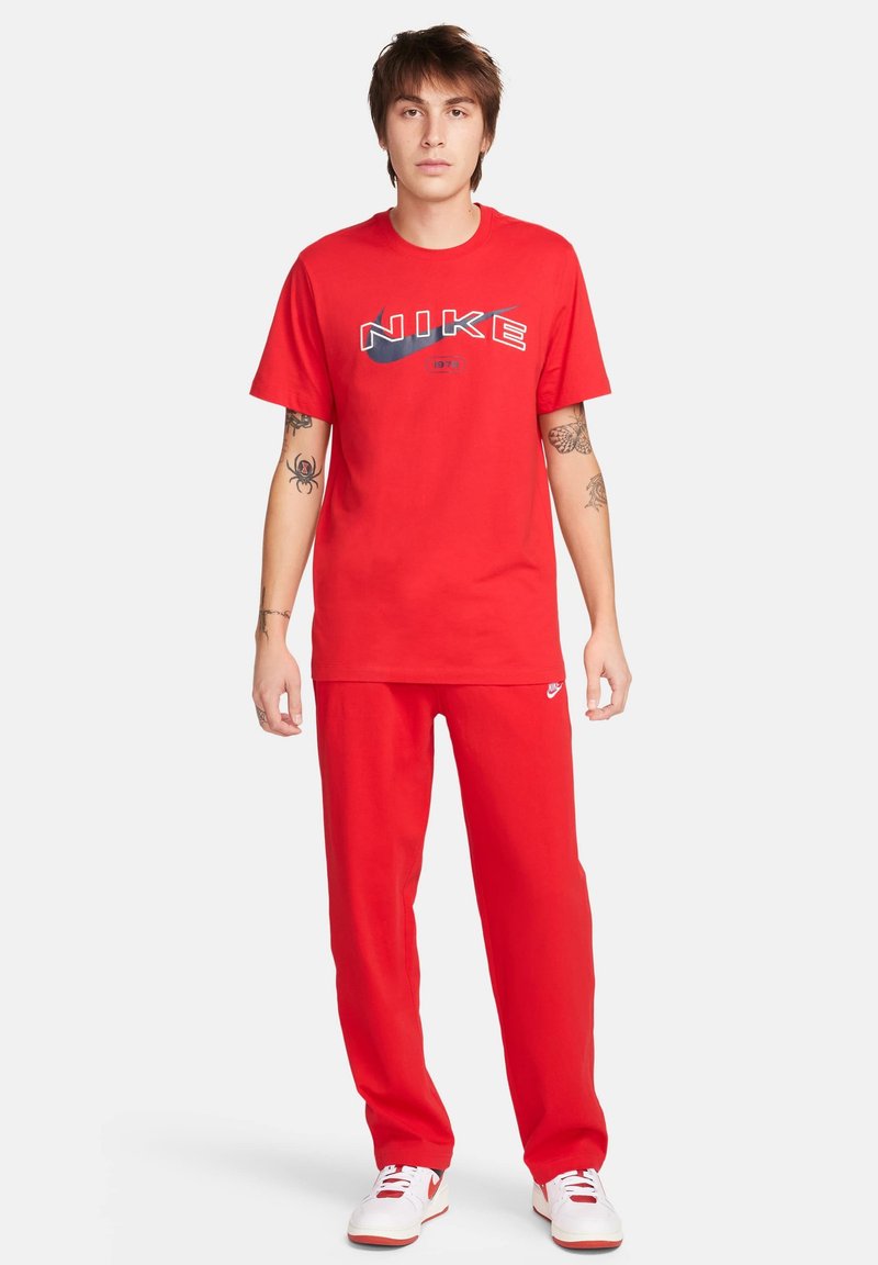 Czerwona koszulka Nike z grafiką logo, zestawiona z dopasowanymi czerwonymi spodniami. Zakończona białymi trampkami. Wykonana z miękkiego materiału o luźnym kroju.