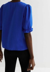 Blouse à manches bouffantes bleu royal avec un col rond, tissu texturé et volants élastiques sur les manches ; associée à un pantalon noir ajusté.