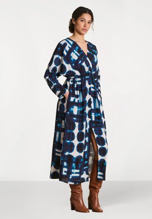 Femme portant une robe midi à manches longues bleu et blanc à motifs, avec un décolleté en V profond et des bottes à talons marron, debout les mains dans les poches.