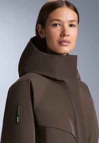 Hnedá nepremokavá bunda s vysokým golierom, hladkou textúrou a minimalistickým dizajnom. Má vrecko na ľavom rukáve a prešité detaily.