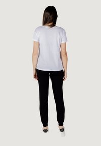 T-shirt bianca in cotone con maniche corte, indossata con pantaloni neri. La maglietta ha un collo rotondo e una vestibilità casual, visibile da dietro.