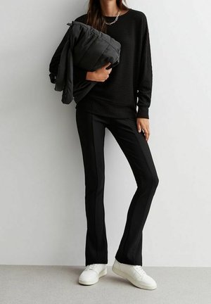 Schwarzer gerippter Pullover mit langen Ärmeln, kombiniert mit schmal geschnittenen schwarzen Hosen mit seitlichen Streifen. Weiße Sneaker vervollständigen das Outfit.