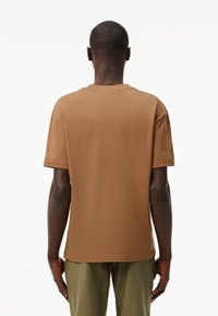 T-shirt a maniche corte marrone, texture liscia, collo rotondo, taglio dritto, indossato con pantaloni verde oliva, vista posteriore, senza motivi o accenti.