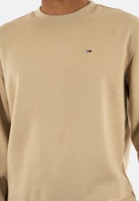 Felpa beige realizzata in tessuto morbido, con collo a girocollo a coste e un piccolo logo ricamato in rosso, bianco e blu sul petto.