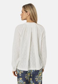 Hvid bluse med lange ærmer og struktureret mønster, samlet bærestykke og draperet rygdetalje, parret med bukser i blå blomsterprint.