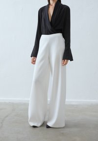 Blusa nera avvolgente con maniche lunghe a campana, abbinata a pantaloni bianchi a vita alta e larghi e décolleté neri a punta. Texture liscia.