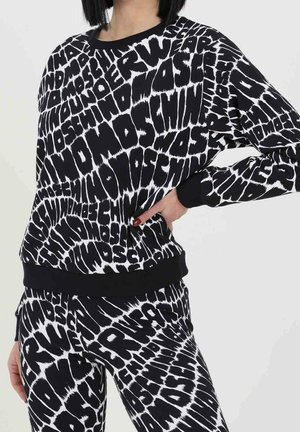 Nainen, jolla on yllään mustavalkoinen Moschino-kuvioitu collegepaita ja housut, joissa on rohkea, vääristynyt tekstisuunnittelu.