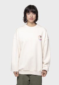 Κρέμα χρώματος oversized φούτερ με στρογγυλή λαιμόκοψη, κατεβασμένους ώμους και ένα μικρό, πολύχρωμο λογότυπο με παλμό στο στήθος.