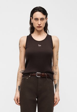 Persoană cu păr lung și întunecat, purtând un top fără mâneci maro închis cu un mic logo de animal și pantaloni asortați cu o curea împletită.