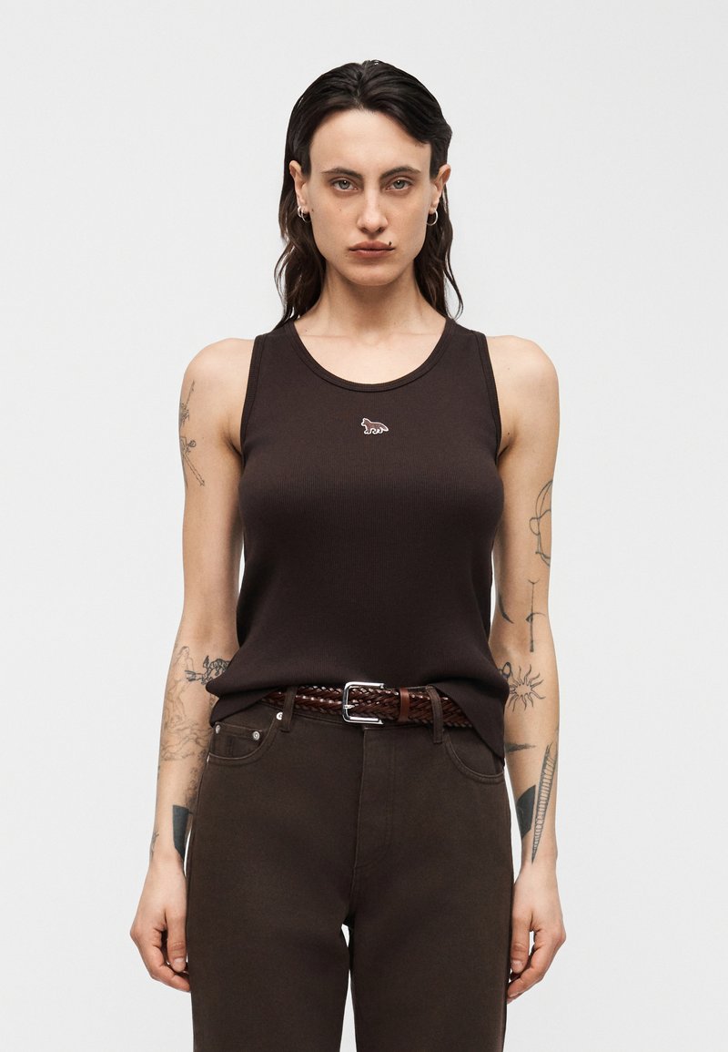 Άτομο με μακριά σκούρα μαλλιά που φοράει σκούρο καφέ αμάνικο τοπ με μικρό λογότυπο ζώου και παντελόνι που ταιριάζει με πλεκτή ζώνη.