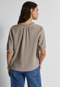 Beige, strukturierte Bluse mit einem geschlitzten Hals, kurzen aufgerollten Ärmeln und einem fließenden Rückendesign, kombiniert mit blauen Denim-Jeans.