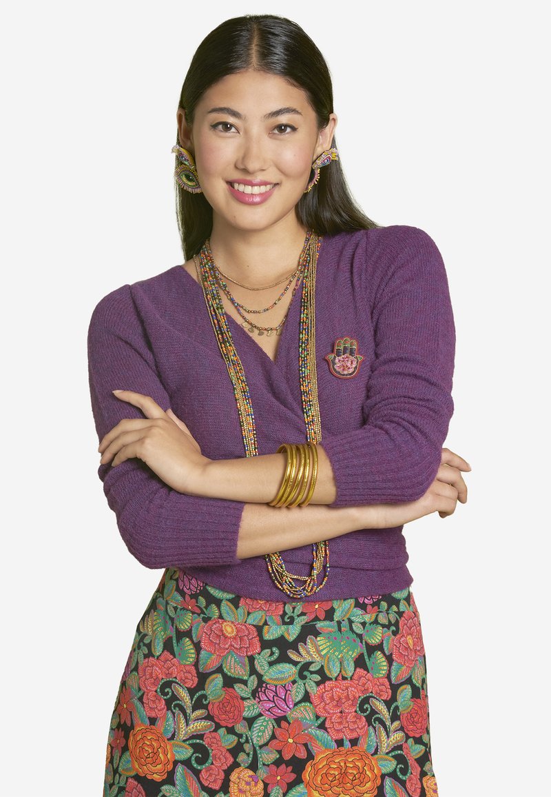 Purple knit sweater with a decorative hand pin, layered gold necklaces, and chunky gold bangles, paired with a floral-patterned black skirt.