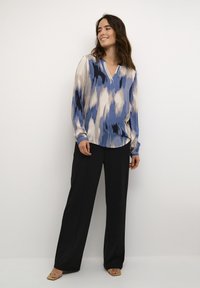 Langärmlige Bluse in Blau und Creme mit Muster und V-Ausschnitt, kombiniert mit schwarzen weiten Hosen und beigen Sandalen an einem Model.