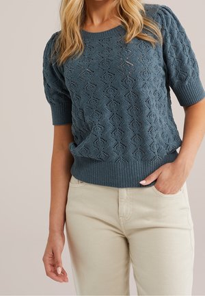 Femme portant un pull bleu texturé à manches courtes et un pantalon beige clair, une main dans la poche et des cheveux blonds visibles.