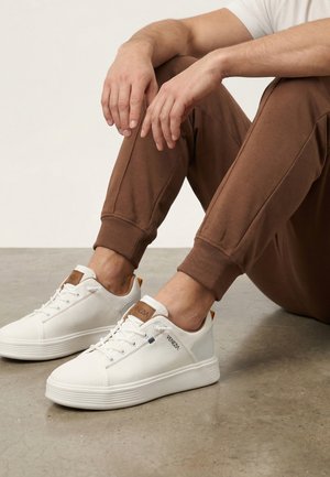 Osoba sedí na podlaze, má na sobě hnědé tepláky a bílé tenisky Venezia s texturovanou látkou a hnědými koženými záložkami na patě.