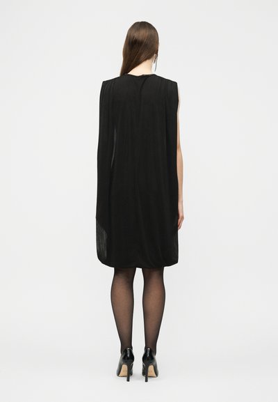 Hosszú barna hajú nő, fekete ujjatlan, térdig érő ruhát, áttetsző fekete harisnyát és fekete magassarkút visel, háttal áll fehér háttér előtt.