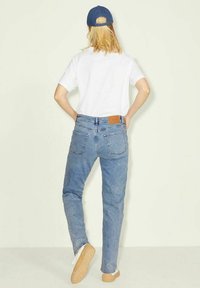 Jeans de ganga azul claro com um corte relaxado, apresentando um retângulo de couro castanho na parte de trás da cintura e bolsos traseiros clássicos. T-shirt branca e sapatos bege.