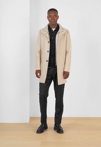 Abrigo de lana beige con frente de botones, cuello y forro gris, llevado sobre una camiseta polo negra y pantalones negros, combinado con zapatos negros.
