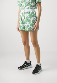 Pantalones cortos blancos con un patrón de hojas verdes, cintura elástica con cordón. Se usan con zapatillas deportivas negras con detalles en verde y calcetines blancos.