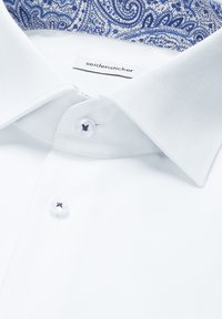 Chemise blanche avec un motif cachemire bleu à l'intérieur du col, dotée de boutons blancs avec un fil bleu et d'une étiquette "seidensticker" visible.