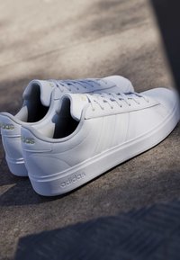 Par hvide Adidas sneakers med snørebånd på betonoverflade, der viser sidestriber og Adidas-logo på sål og hæl.