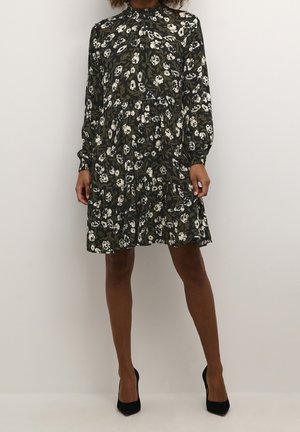 Kvinde iført en mørk blomsterprint langærmet kjole med lagdelt nederdel og sorte spidse hæle, stående mod en ensfarvet lys baggrund.