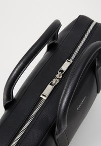 Schwarze Lederhandtasche mit silbernem Reißverschluss, doppelten Griffen und einer strukturierten Oberfläche. Mit einem Calvin Klein-Logo an der Seite.