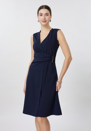 Γυναίκα που φοράει αμάνικο μπλε σκούρο φόρεμα μέχρι το γόνατο με περιτύλιγμα και πλευρικό κούμπωμα, στέκεται μπροστά σε απλό φωτεινό φόντο.