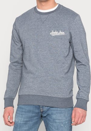 Muž v šedé mikině Jack & Jones s žebrovanými manžetami a lemem, doplněné modrými džíny.