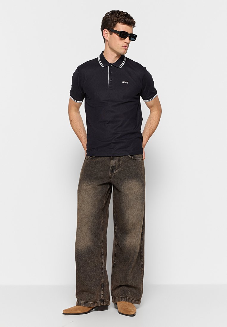 Marineblaues Polohemd mit weiß gestreiftem Kragen, kombiniert mit lockeren braunen Denim-Jeans. Das Modell trägt außerdem schwarze Sonnenbrillen und beigefarbene Schuhe.