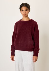 Moteris garbanotais plaukais, dėvinti auksinius lankinius auskarus, bordo vilnonį megztinį ir šviesiai pilkas kelnes, stovinti priešais paprastą foną.