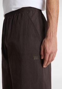 Dark brown pants featuring a textured fabric, elastic waistband, side pockets, and a small embroidered "VANS" logo on the lower leg.