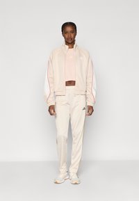 Φωτεινή μπεζ φόρμα που περιλαμβάνει ζακέτα με φερμουάρ και λευκές λεπτομέρειες με κόκκινες ρίγες, συνδυασμένη με αντίστοιχο παντελόνι. Το λογότυπο της Adidas είναι ορατό και στα δύο.