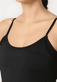 Mujer vestida con una blusa negra sin mangas de tirantes finos, mostrada desde el cuello hasta la parte superior del pecho sobre un fondo neutro.