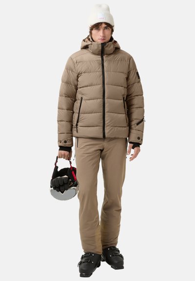 Beige puffer jacket with a hood, zippered pockets, and black accents; beige pants; holds a ski helmet; black ski boots; neutral background.