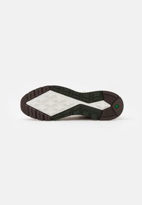 Semelle de chaussure avec bande de roulement en caoutchouc marron et noir, motif de grip blanc en forme de croix et logo hexagonal vert près du talon.
