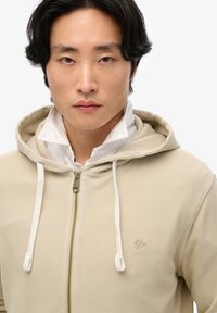 Sudadera con cremallera en color beige, con capucha ajustable y tejido texturizado, que cuenta con un pequeño logo bordado en el pecho. Cuello blanco visible por debajo.
