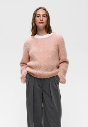 Kvinde iført en blød lyserød fluffy sweater og grå plisserede bukser, stående med hænderne i lommerne mod en ensfarvet lys baggrund.