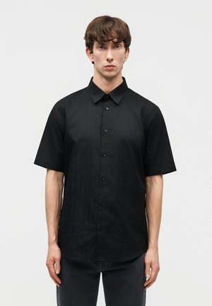 Νέος άνδρας με ανοιχτόχρωμο δέρμα και καστανά μαλλιά, φορώντας κοντομάνικο μαύρο πουκάμισο με κουμπιά και σκούρο παντελόνι, στέκεται μπροστά σε ένα απλό λευκό φόντο.