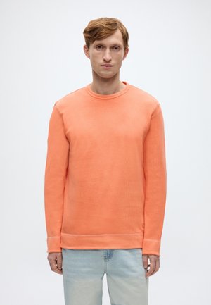 Pomarańczowa prążkowana bluza z okrągłym dekoltem, długimi rękawami i prostym dołem, zestawiona z jasnoniebieskimi dżinsami, na jednolitym tle.