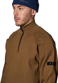 Brauner Pullover mit Reißverschluss und gerippter Textur, ausgestattet mit einem schwarzen Stoffpatch mit "AT" am linken Arm und einem hohen Kragen.