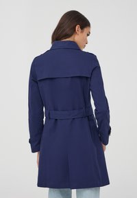 Trench coat blu navy con vita regolabile, grande colletto e piega posteriore. Realizzato in tessuto liscio, presenta maniche con bottone e vestibilità sartoriale.