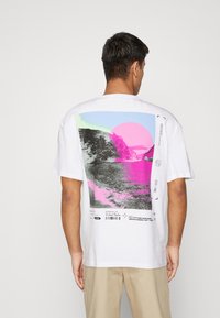 Weißes Baumwoll-T-Shirt mit einem grafischen Druck auf der Rückseite in rosa und blauen Farbtönen, das eine Küstenszene und geometrische Formen darstellt.