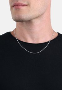 Primo piano di una persona che indossa una sottile collana d'argento e una maglietta nera a girocollo, mostrando la mascella e il collo su uno sfondo neutro.