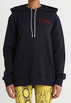 Person trägt einen marineblauen Hoodie mit "CREW" in roten Buchstaben und weißen Kordeln, kombiniert mit gelber Hose im Schlangenledermuster.